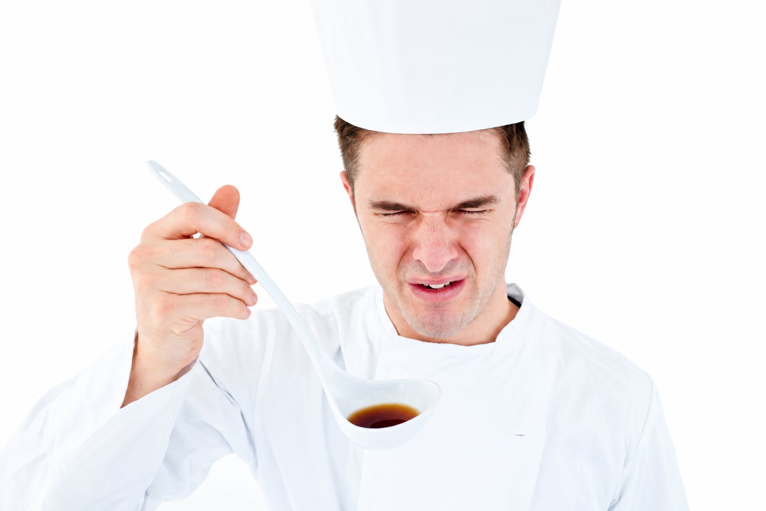 Un cuisinier en veste et toque blanches fait une grimace en goûtant un liquide brun avec une louche, sur fond blanc.