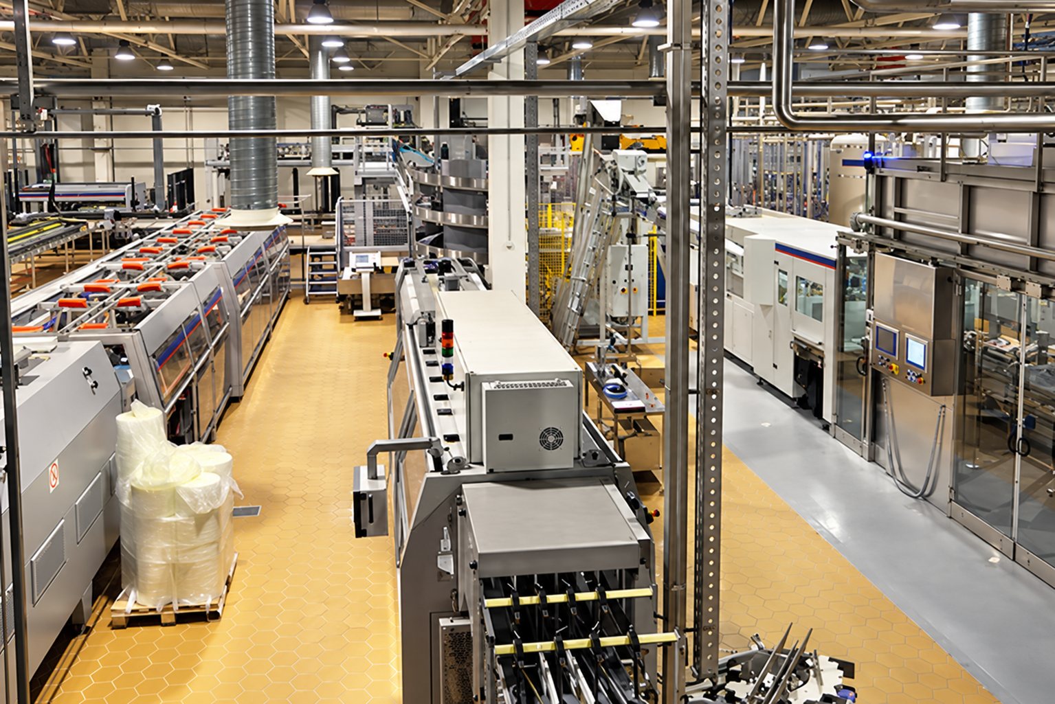 Vue plongeante sur une grande usine équipée de machines de production automatisées, de tuyauteries et de convoyeurs sur un sol clair.